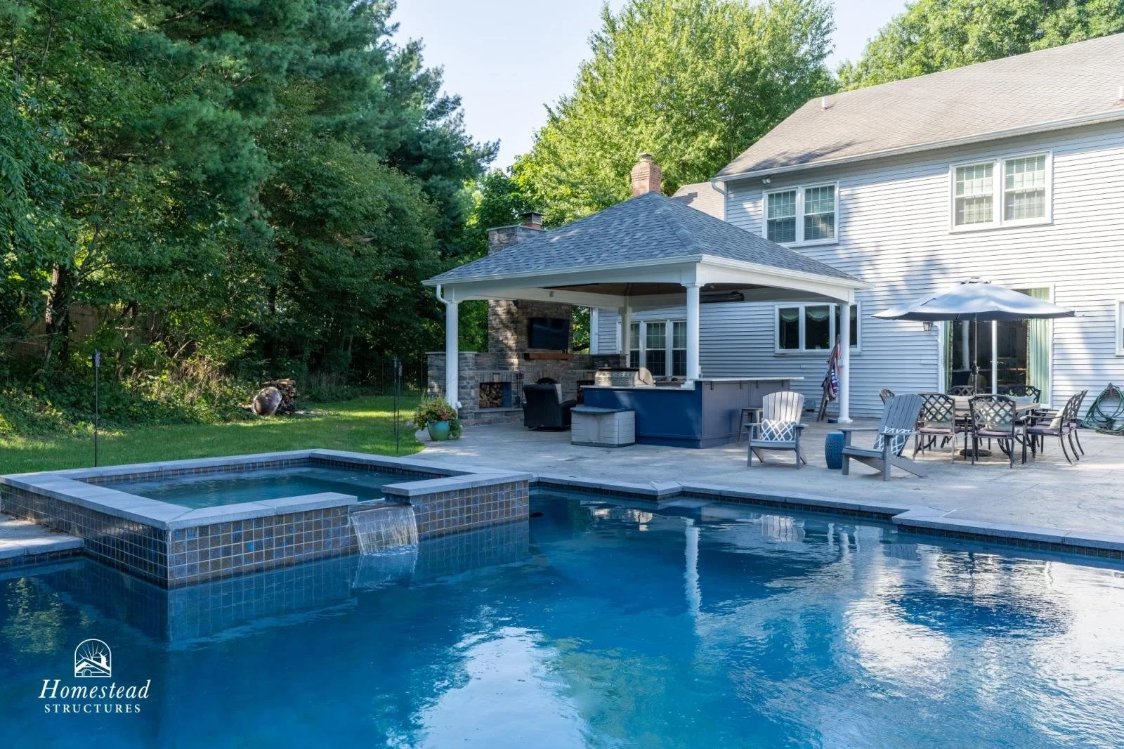 Backyard with pool and hot tub, patio with outdoor furniture, umbrella, and built-in outdoor fireplace, surrounded by trees and a two-story house.