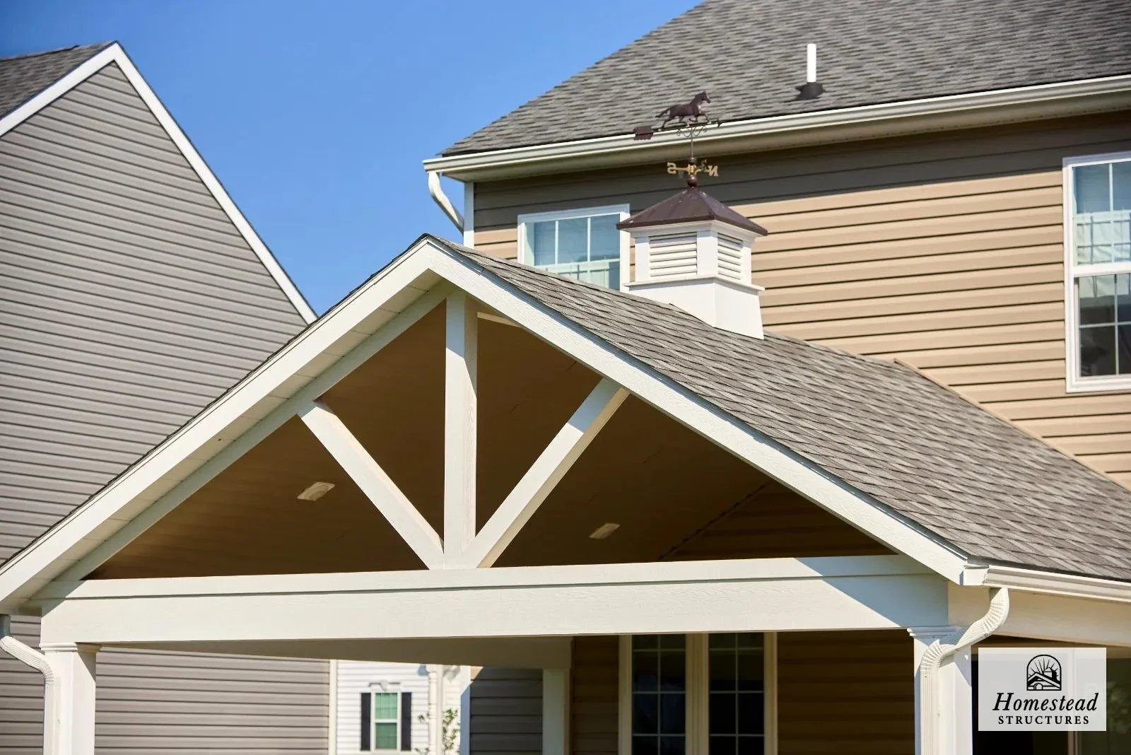 A residential house with beige siding, a gable roof with shingles, and a covered porch area with wooden beams painted white. There's a small chimney and a weather vane on the roof.
