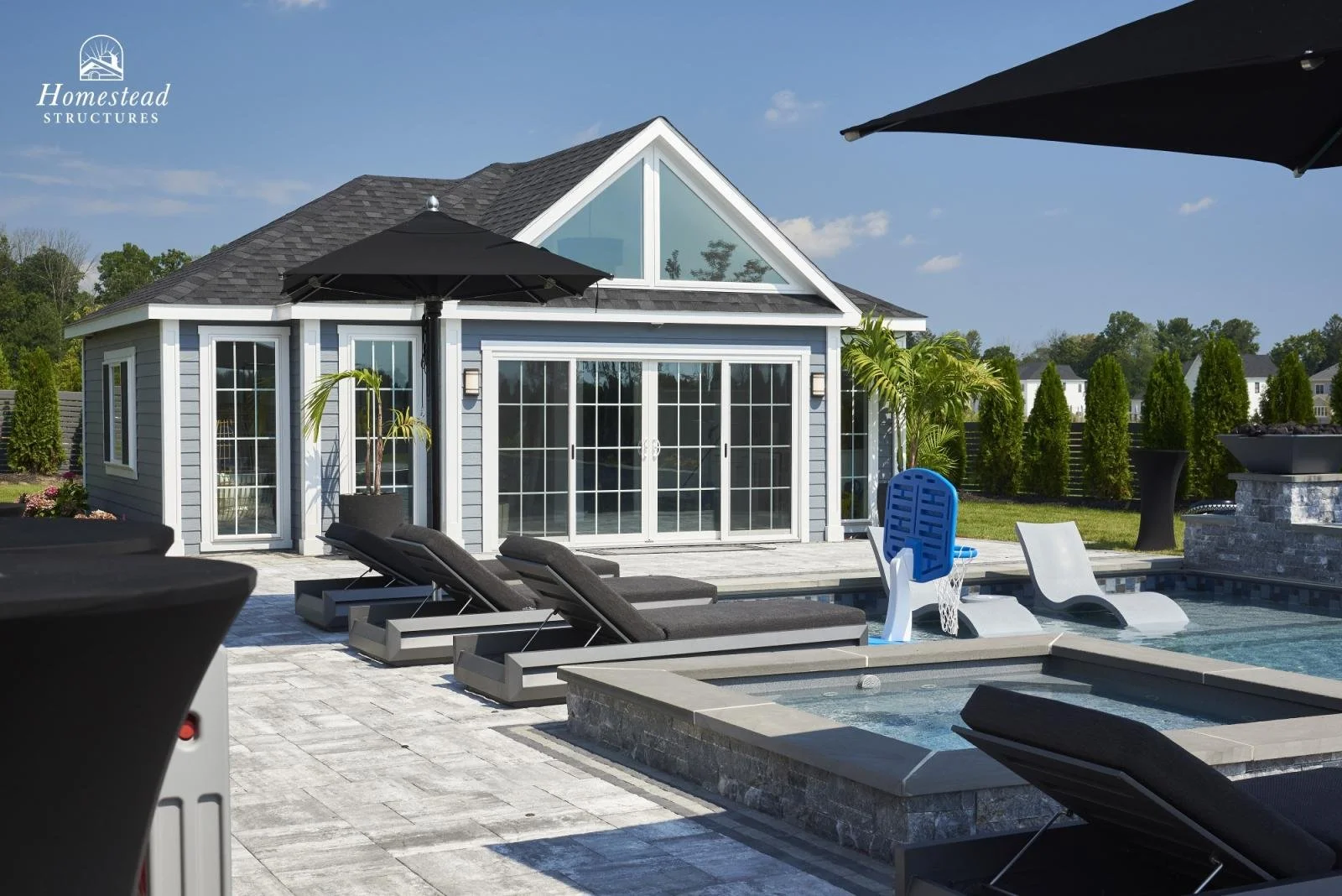 A backyard with a swimming pool and a modern house with large glass sliding doors, surrounded by greenery and tall trees, with black umbrellas and lounge chairs.