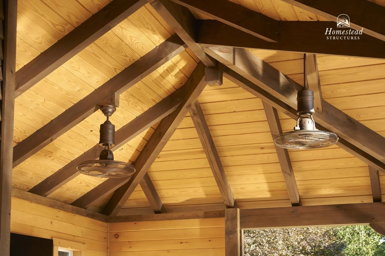 Interior view of a wooden porch ceiling with exposed beams, two ceiling fans, and sunlight coming through an open area at the bottom.