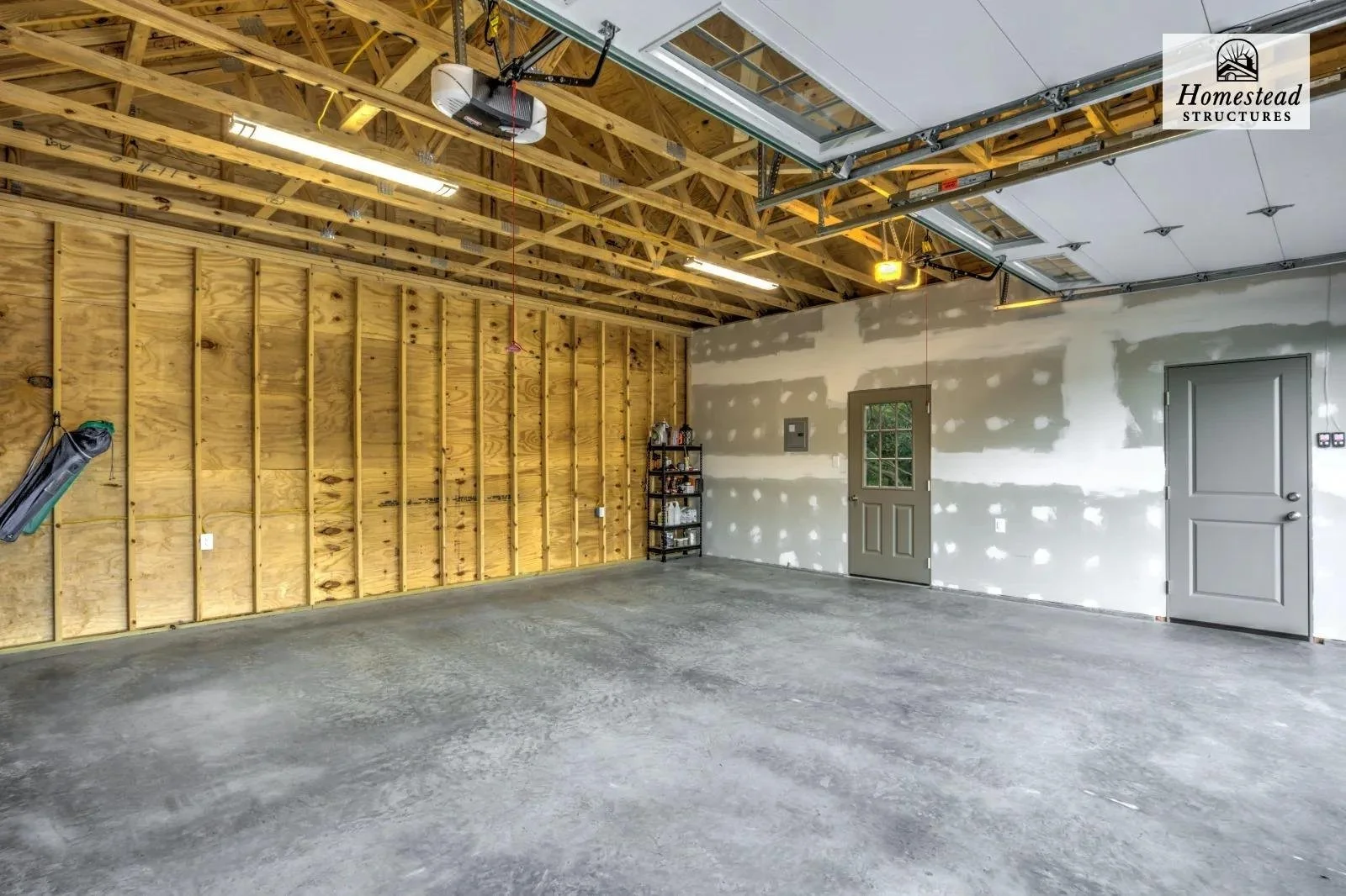 Empty garage with unfinished walls, exposed wooden ceiling structure, concrete floor, two gray doors, garage door opener, and a small black shelf with supplies.