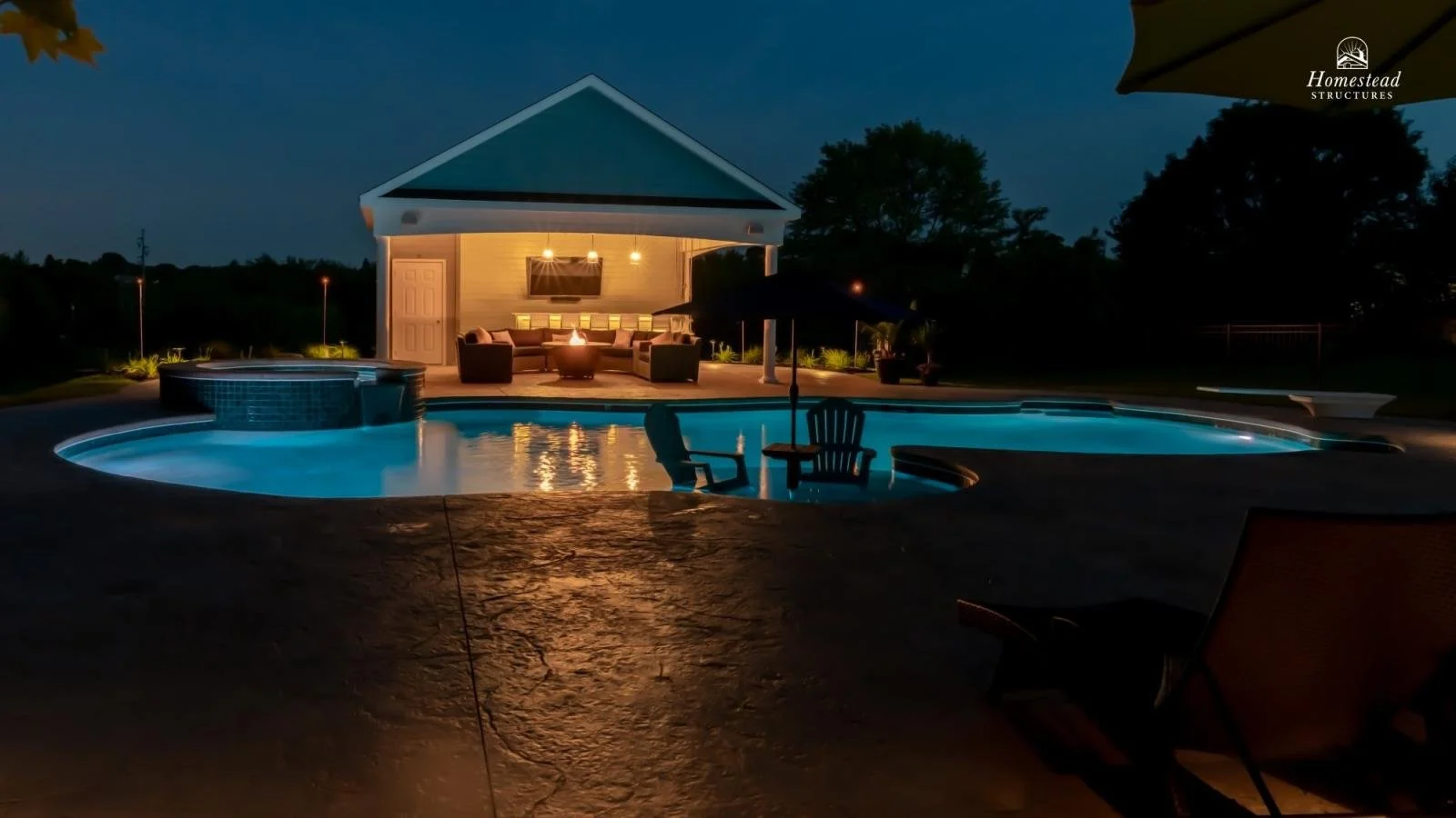 Nighttime view of a backyard with a lit pool, hot tub, and a covered patio area with outdoor seating, under string lights, with trees in the background.