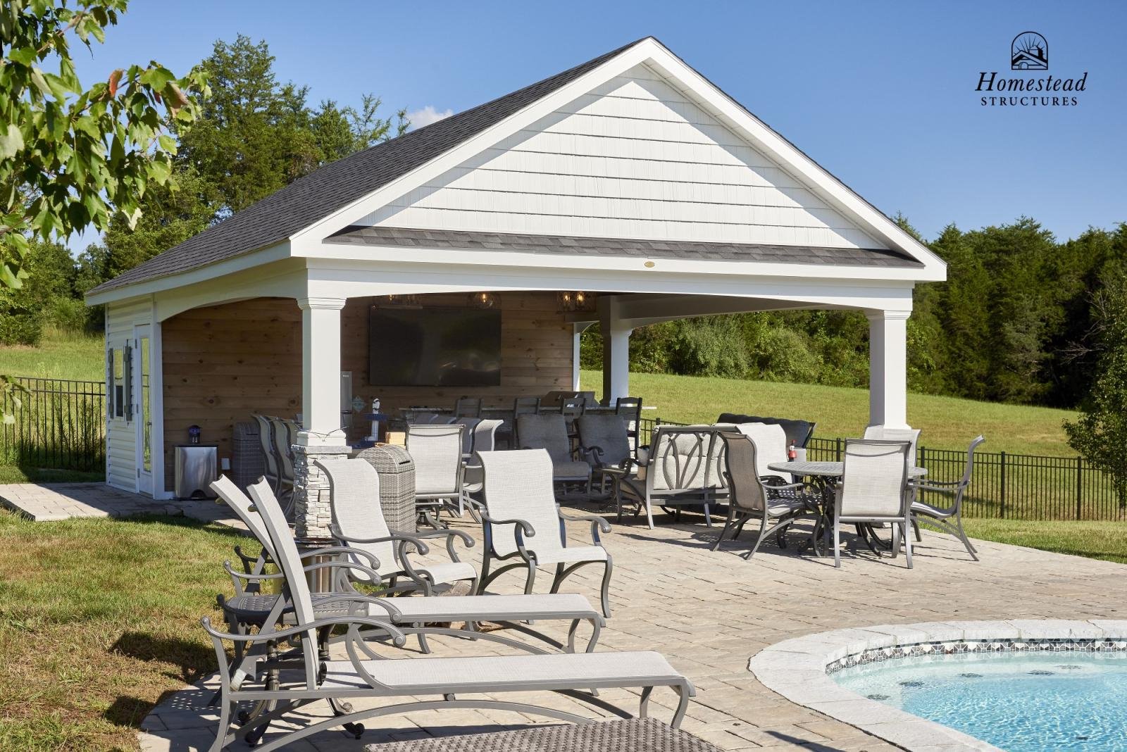 Backyard patio with lounge chairs, a swimming pool, and a covered outdoor living area with a large TV and seating, surrounded by green grass, trees, and a black metal fence. The image features a backyard with a pool and outdoor furniture with a logo 