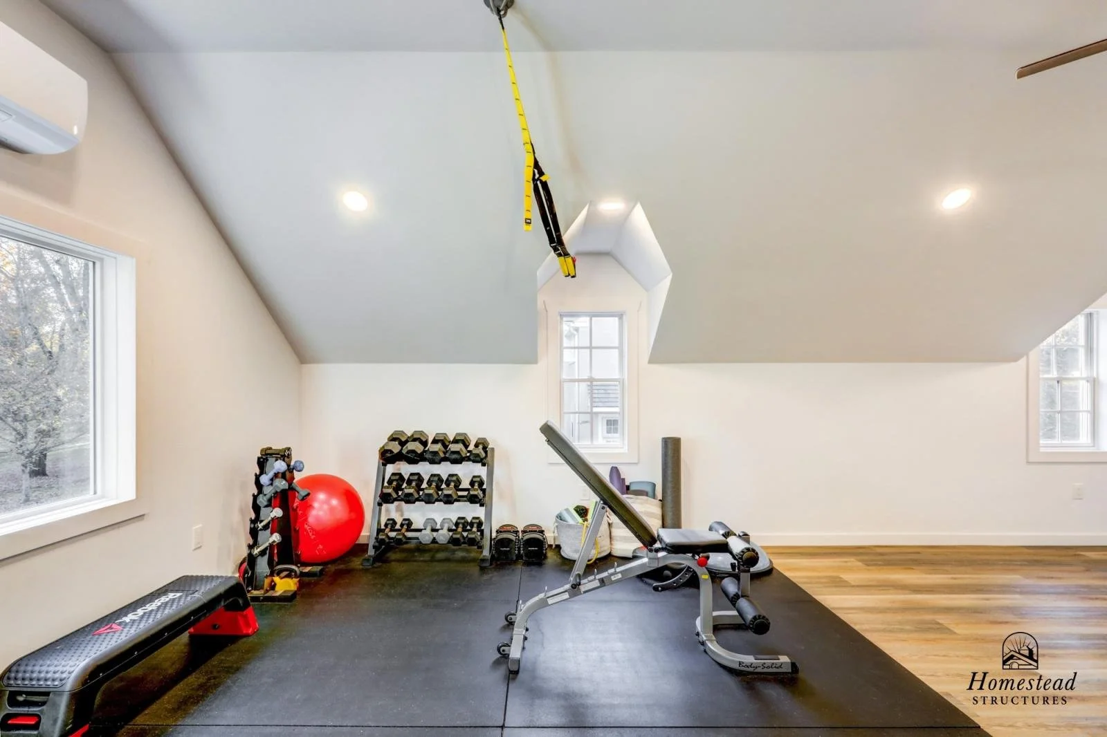 Home gym with workout equipment including a weight bench, dumbbell rack, stability ball, exercise mats, and a suspension trainer hanging from the ceiling in a room with white walls and hardwood floors.