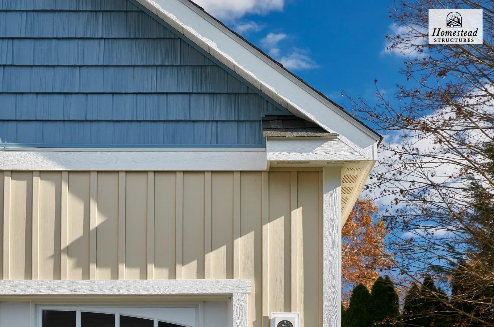 Close-up of the corner of a house with beige siding, a white trim, and blue shingle siding under the roof, with a blue sky and trees with fall foliage in the background.