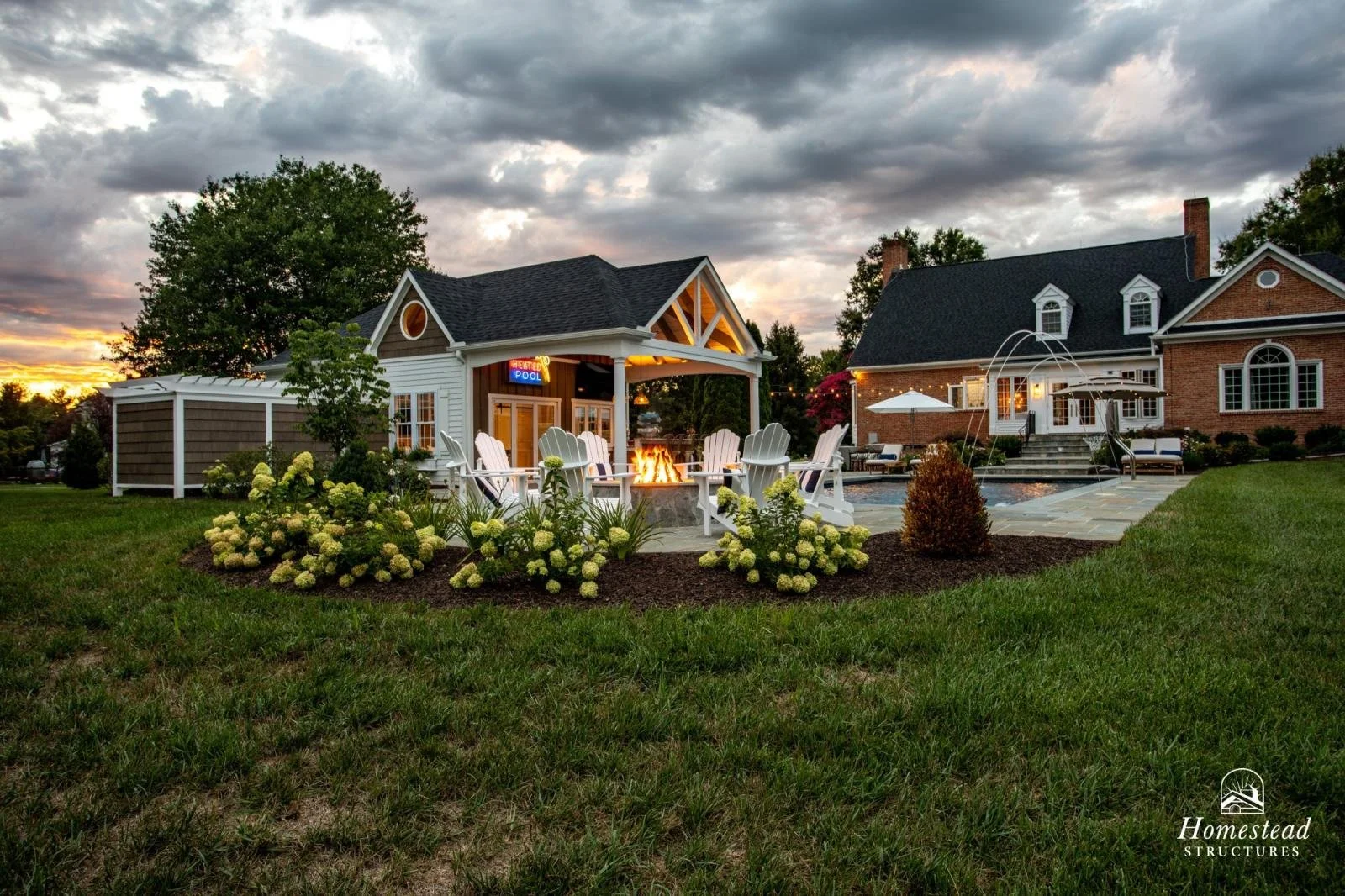 A backyard with a fire pit, Adirondack chairs, a swimming pool, and two houses under a cloudy sunset sky.