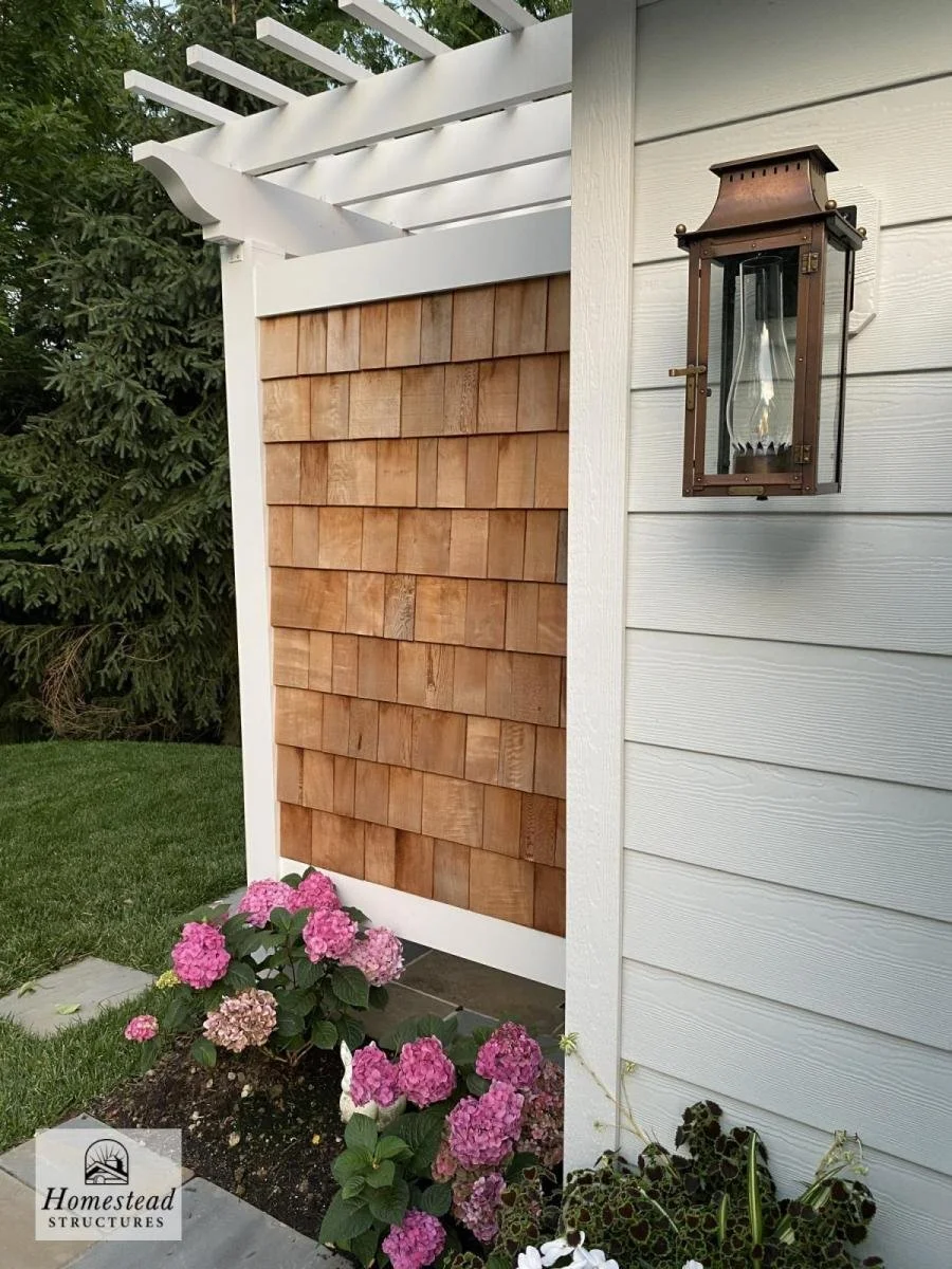 A section of a house exterior featuring a white wooden pergola, a flower bed with pink and purple hydrangeas, a wall-mounted lantern-style light fixture, and part of a white siding wall.
