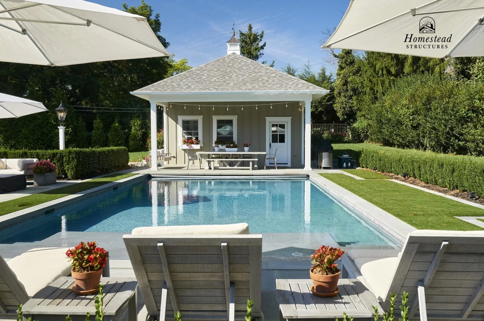 A backyard with a swimming pool, surrounded by lounge chairs and umbrellas, and a pool house with outdoor seating and potted plants.