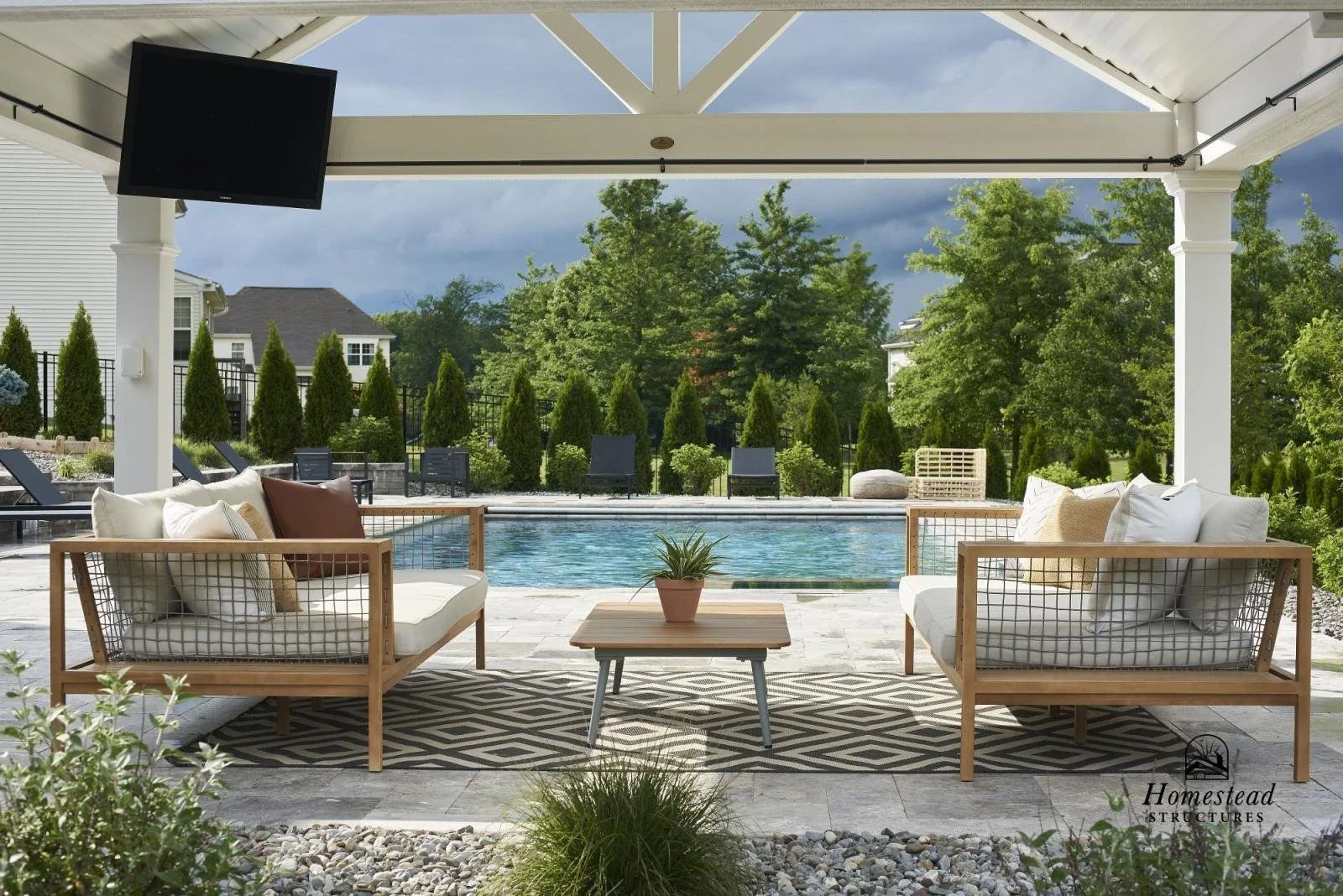 Outdoor patio with two wooden framed couches with neutral cushions, a small wooden coffee table with a plant, and a swimming pool in the background surrounded by greenery and trees.