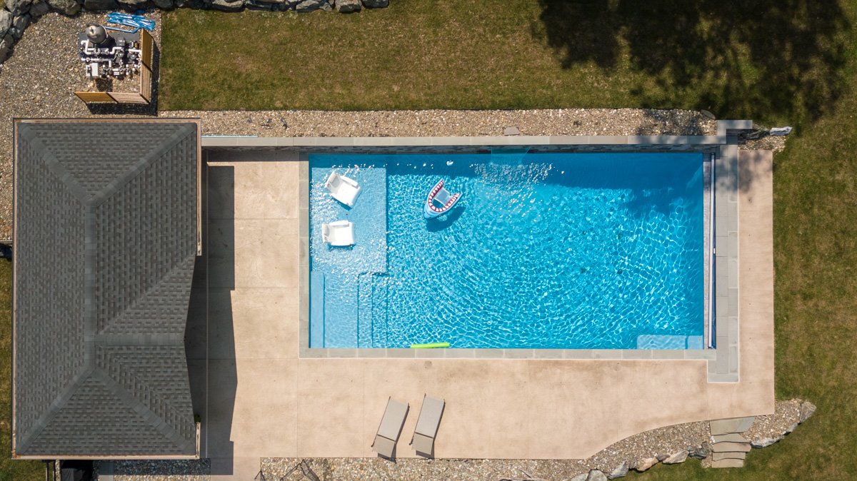 A top-down view of a backyard swimming pool with two lounge chairs, a float, and water toys, surrounded by a concrete patio, grass, and a small rock garden.