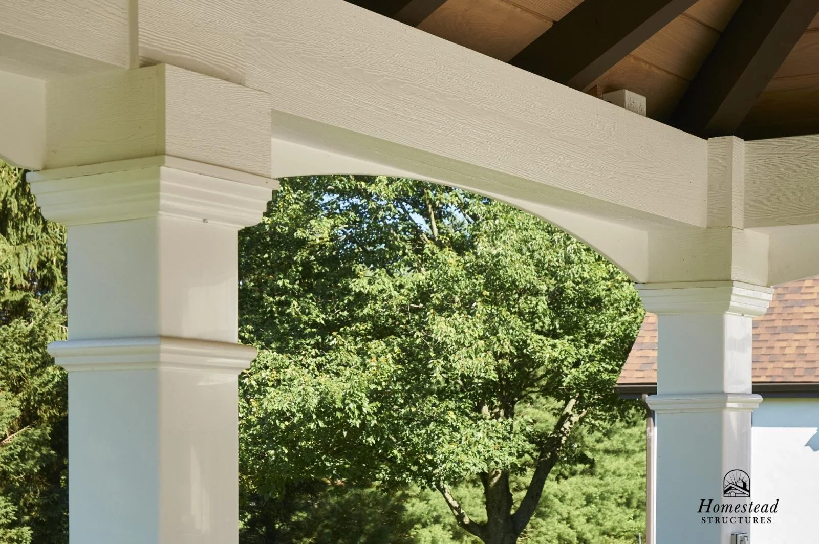Close-up view of a porch with cream-colored columns and decorative crown molding, with green trees and a house roof in the background.