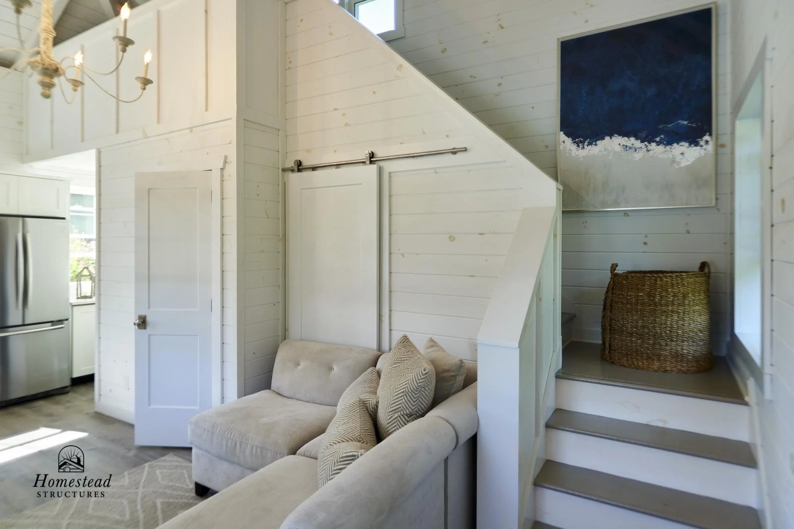 Interior of a cozy, white-painted wooden living space with a small beige sofa and decorative pillows, a staircase leading up, a large blue abstract painting on the wall, and a wicker basket on the landing; part of a kitchen with a stainless steel ref