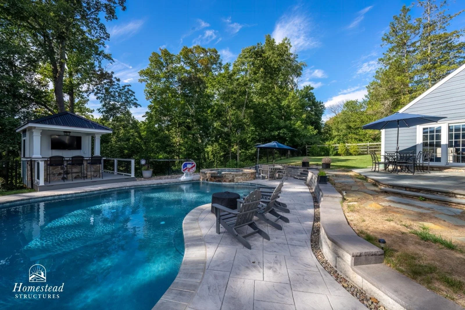A backyard swimming pool with lounge chairs, a hot tub, and a patio with a dining table. There is a small building with a TV on the porch, and greenery with trees in the background.