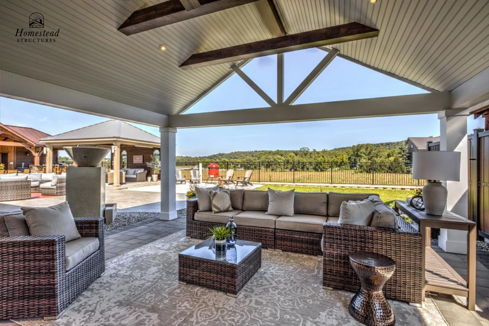 Outdoor covered patio with wicker furniture, cushions, a coffee table, and a lamp in a residential backyard with a view of green fields and trees.