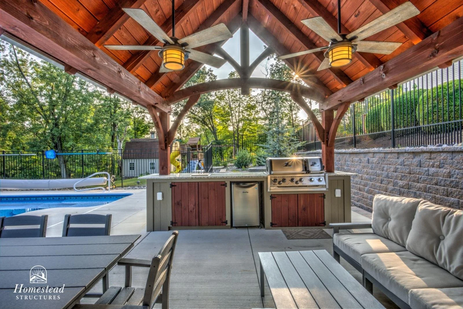 Covered outdoor patio area with a built-in outdoor kitchen featuring a grill, small refrigerator, and wooden cabinet doors. There is a ceiling fan with lights, a seating area with a cushioned sofa, and a dining table with chairs. In the background, t