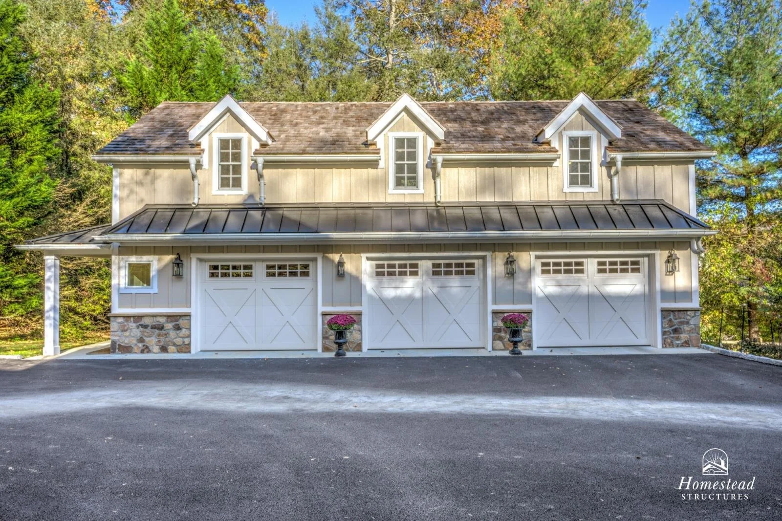 A large residential garage with three white doors, stone accents at the base, and small windows at the top. The house has beige siding, three small gabled dormer windows, and a metal roof. There are trees in the background and two potted plants with 