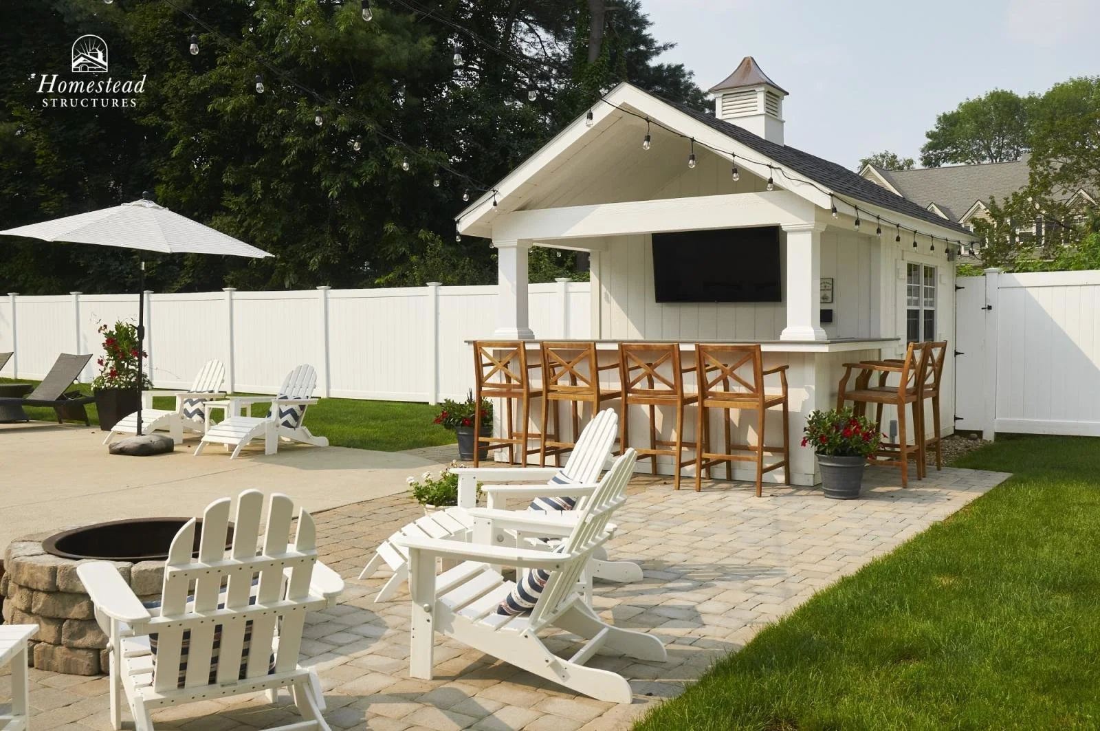 A backyard patio with white Adirondack chairs, a fire pit, a white bar with wooden stools, a white fenced yard, green grass, a white shed with a TV, and outdoor string lights.