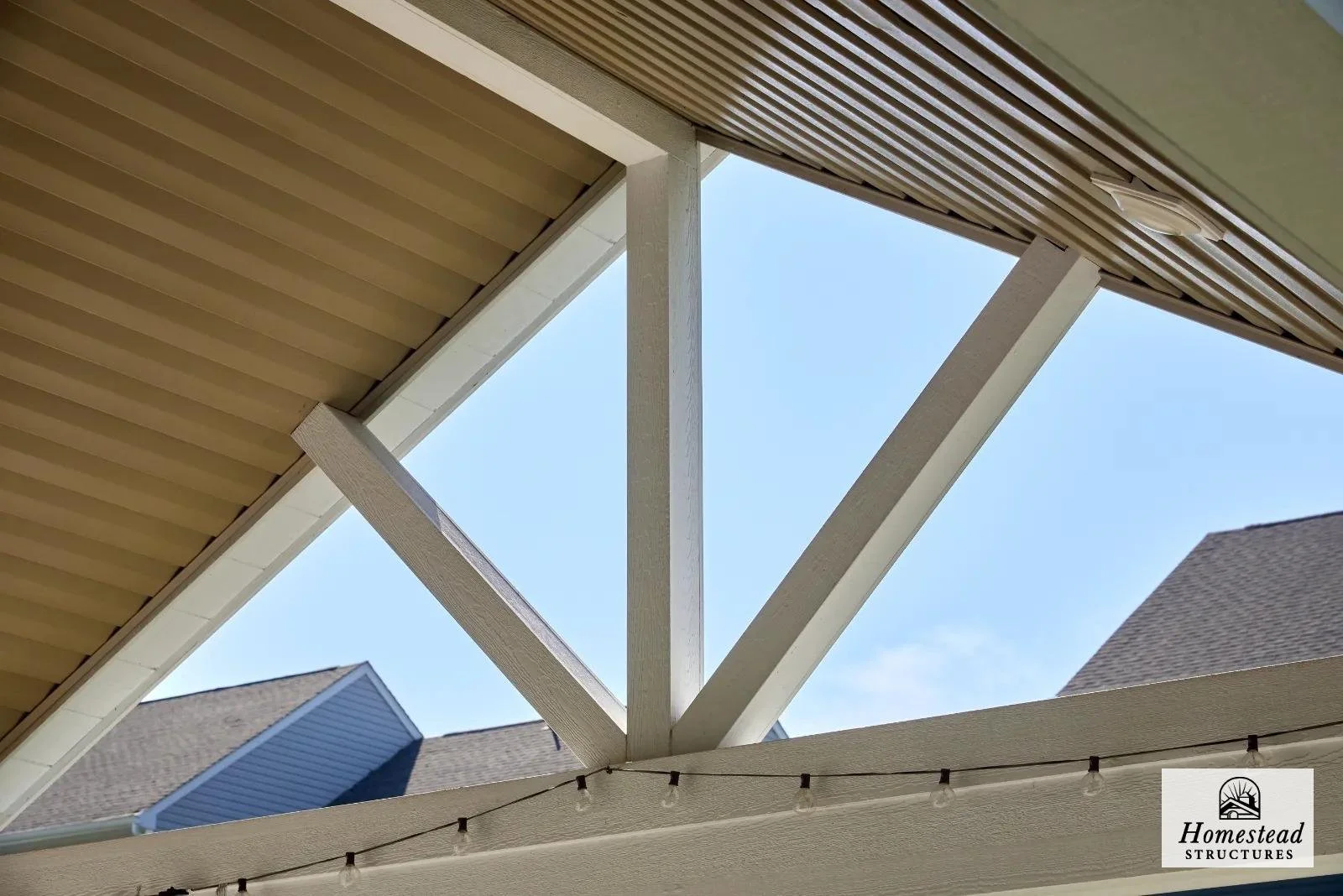 View of house roof overhang with white wooden beams and string lights, blue sky in the background.