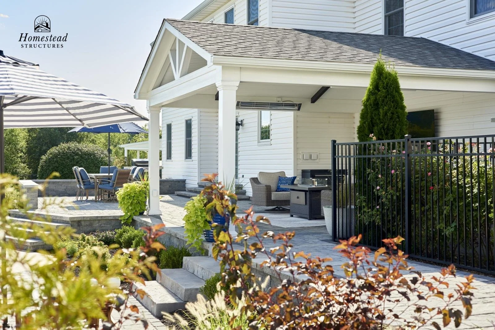 Outdoor patio with chairs, umbrellas, and a grill area, surrounded by plants and a black metal fence.