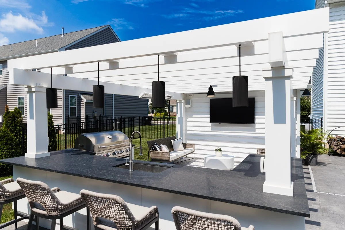 Outdoor patio with a white pergola, black hanging speakers, a flat-screen TV on the white wall, a patio sofa, a white side table, a planter, island with a grill and a sink, barstools, and neighboring houses in the background.