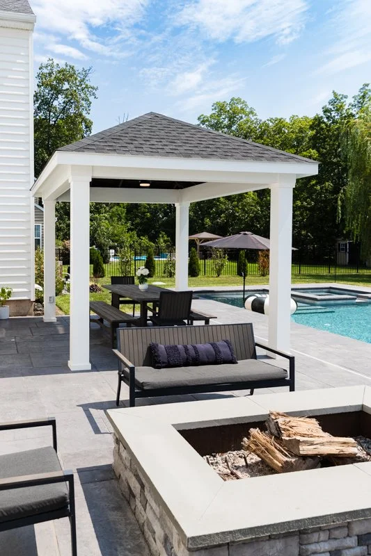 Backyard with pool, gazebo, outdoor seating area, fire pit, and swimming pool under a bright sky.