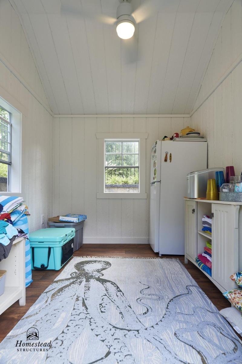 Small room with white wooden walls and ceiling, hardwood floor, two windows, a white refrigerator, a white cabinet with towels, colorful cups, and a rug with an octopus design.