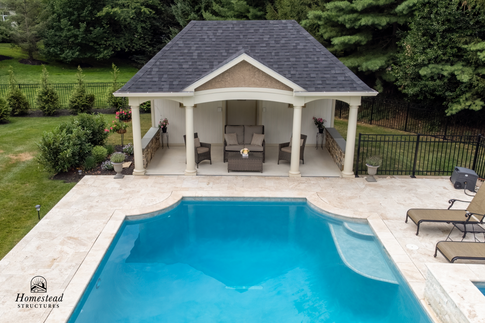 An outdoor swimming pool with a surrounding stone deck, adjacent to a covered patio area with wicker chairs and a small table, set in a green backyard with trees and bushes.