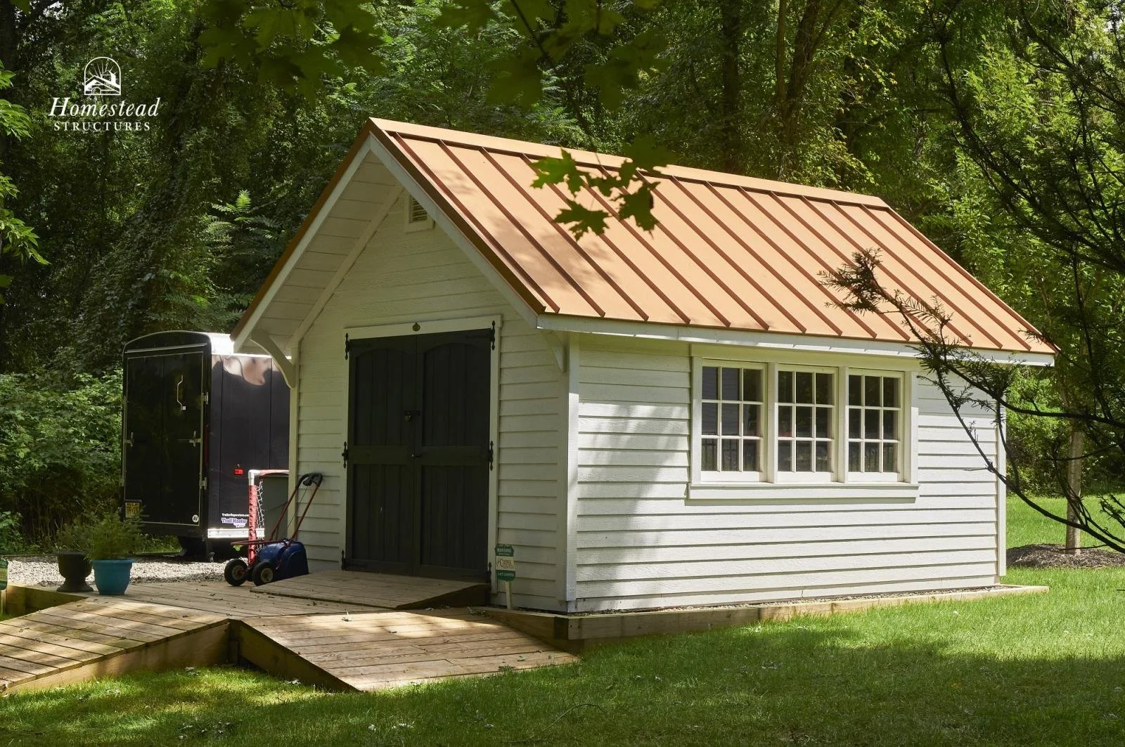 Small white wooden shed with a brown metal roof, double black doors, and a window on the side, located in a grassy backyard with trees.