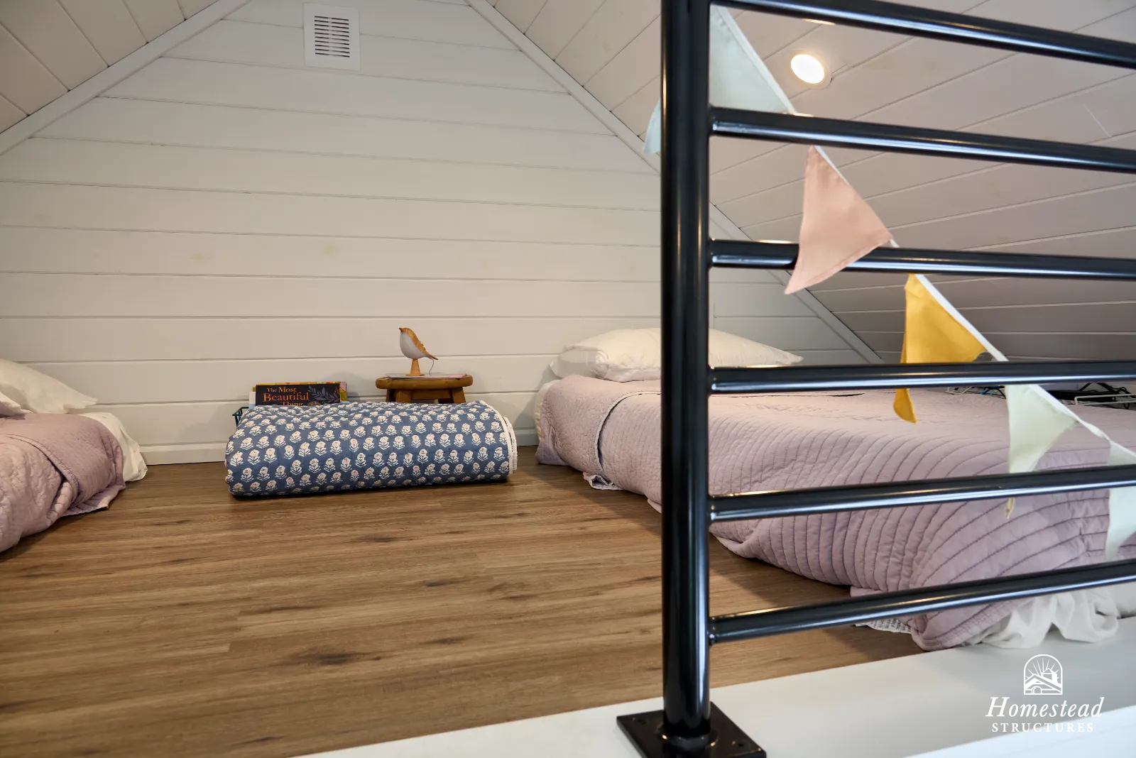 Insulated attic space with three beds on a wooden floor, one with a pink quilt, one with a purple quilt, and the third with a blue patterned quilt. Small side table with books and a decorative bird sculpture. Colorful bunting hanging from black metal