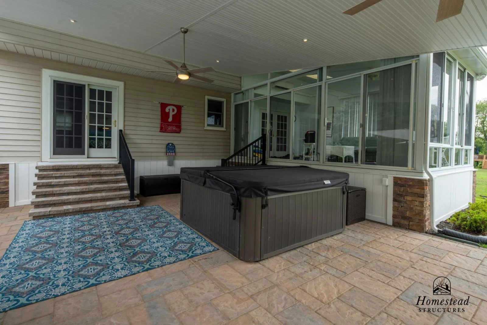 Covered backyard patio with hot tub, brick stairs leading to a glass-enclosed porch, ceiling fan, decorative rug, and Phillies flag.