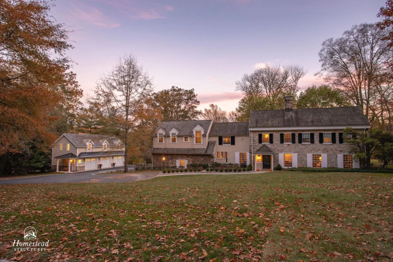 A large house with a stone facade, white shutters, and a chimney, surrounded by trees with autumn foliage and a grassy lawn covered in fallen leaves, during dusk with lights on inside.