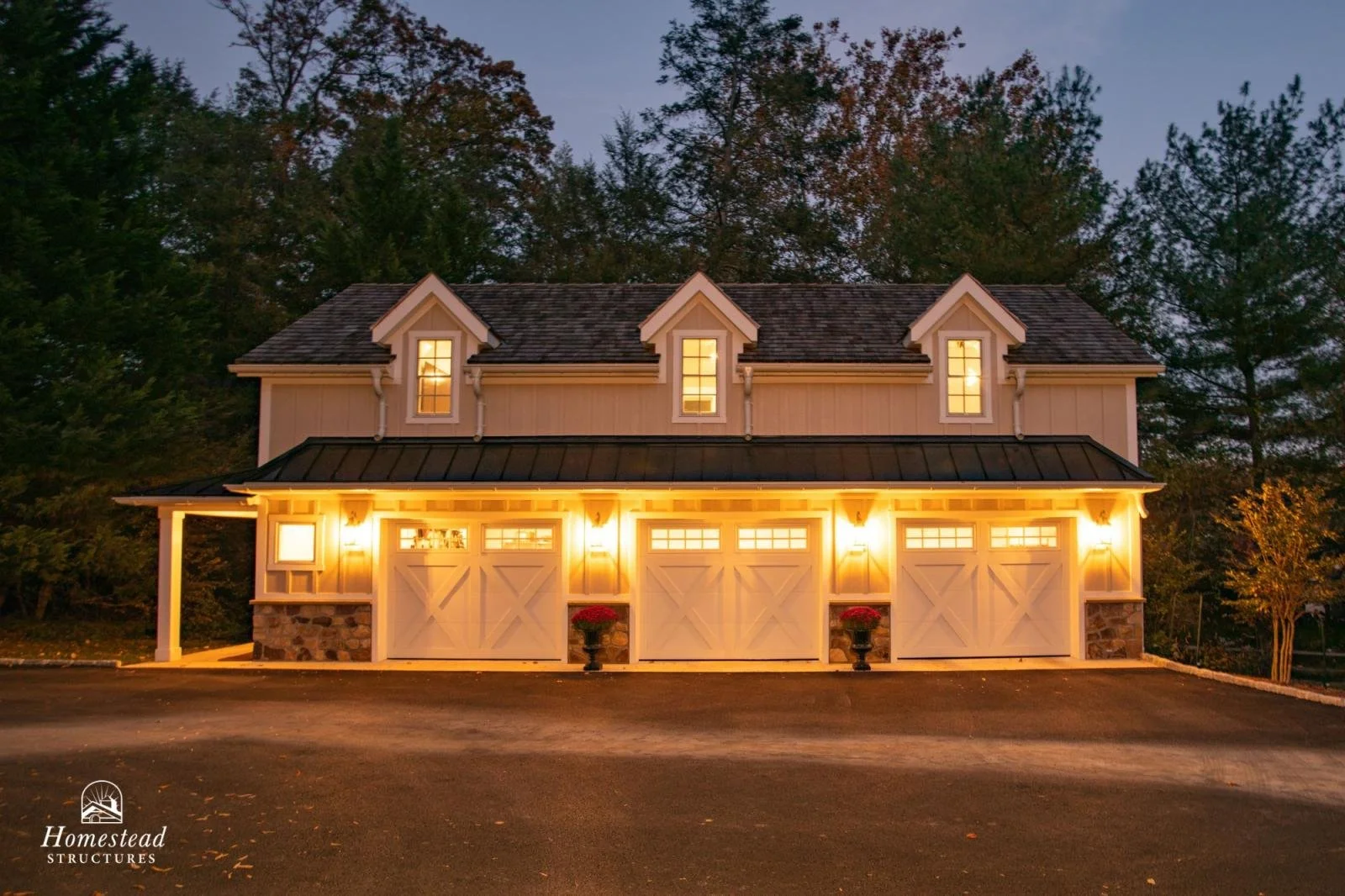 A farmhouse-style garage with three white doors, exterior lights, and small upper windows, surrounded by trees at dusk.