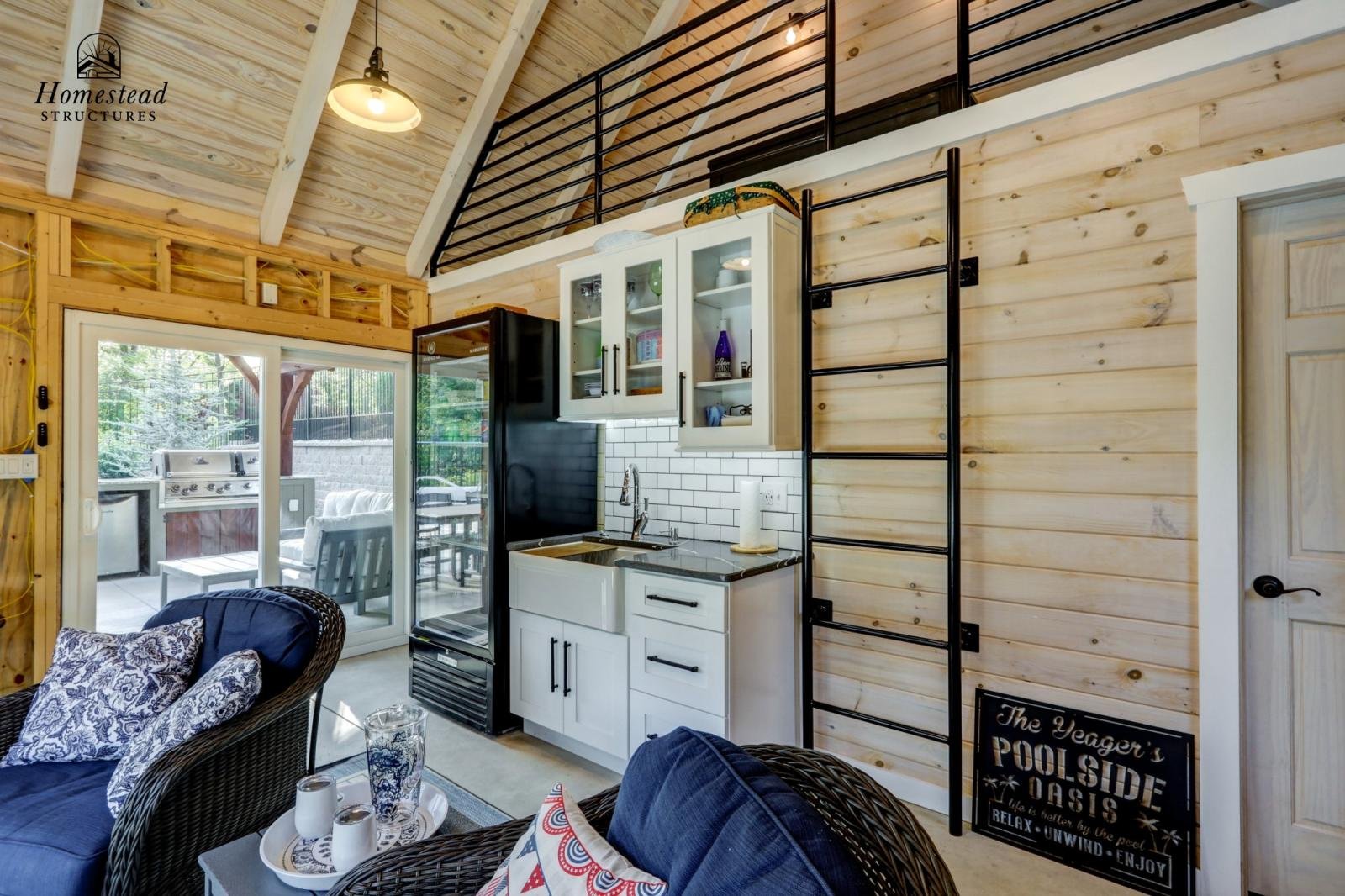 Interior of a rustic-style living space with a small kitchenette, wicker chairs with cushions, and a sliding glass door leading to an outdoor patio with a grill. Light-colored wooden walls and ceiling with exposed beams, black metal ladder, and a sig