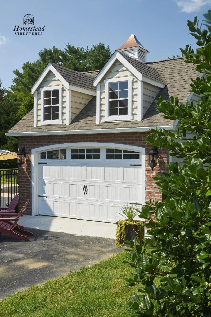 Front view of a house with a white garage door, brick facade, and two windows in dormer structures on the roof, surrounded by green trees and bushes.