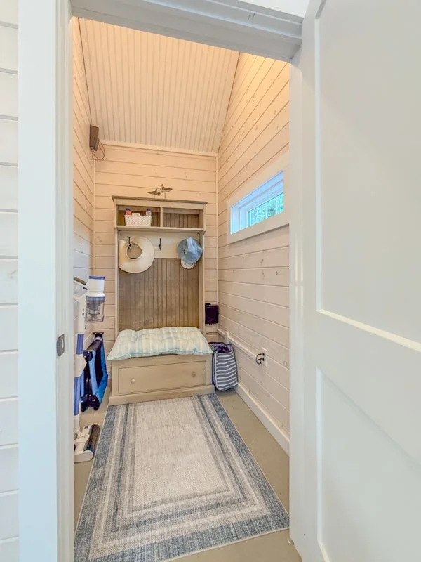 Small entryway with a built-in bench and storage, light-colored wooden walls and ceiling, a small window on the right, and some hanging hats and items on the wall, with a rug on the floor.