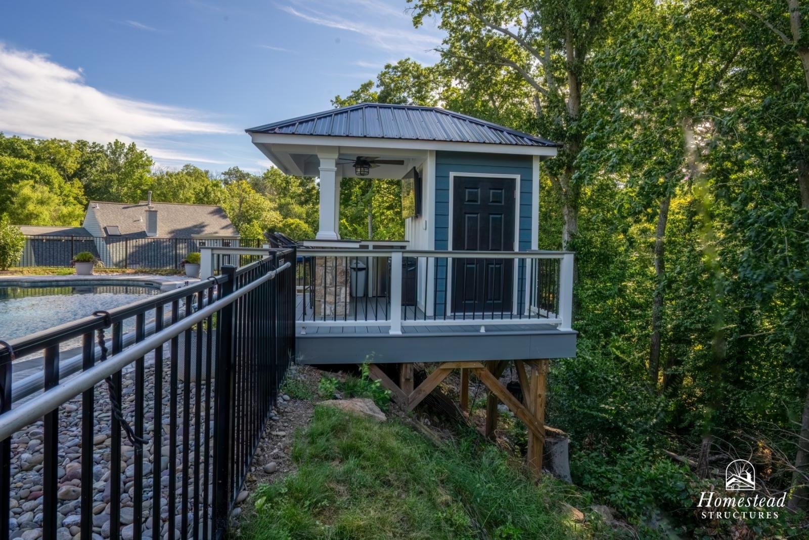 A small, elevated blue structure with a black door, white trim, supported by wooden stilts, and a metal roof, surrounded by dense green trees and a black metal railing, with a swimming pool nearby.