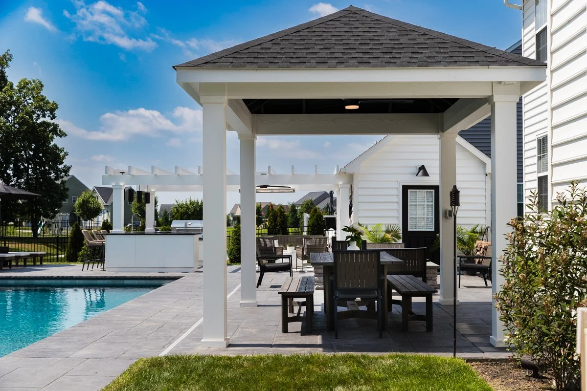 Backyard with pool, outdoor seating area under a white pavilion, and a small white house with black door and windows, sunny sky.