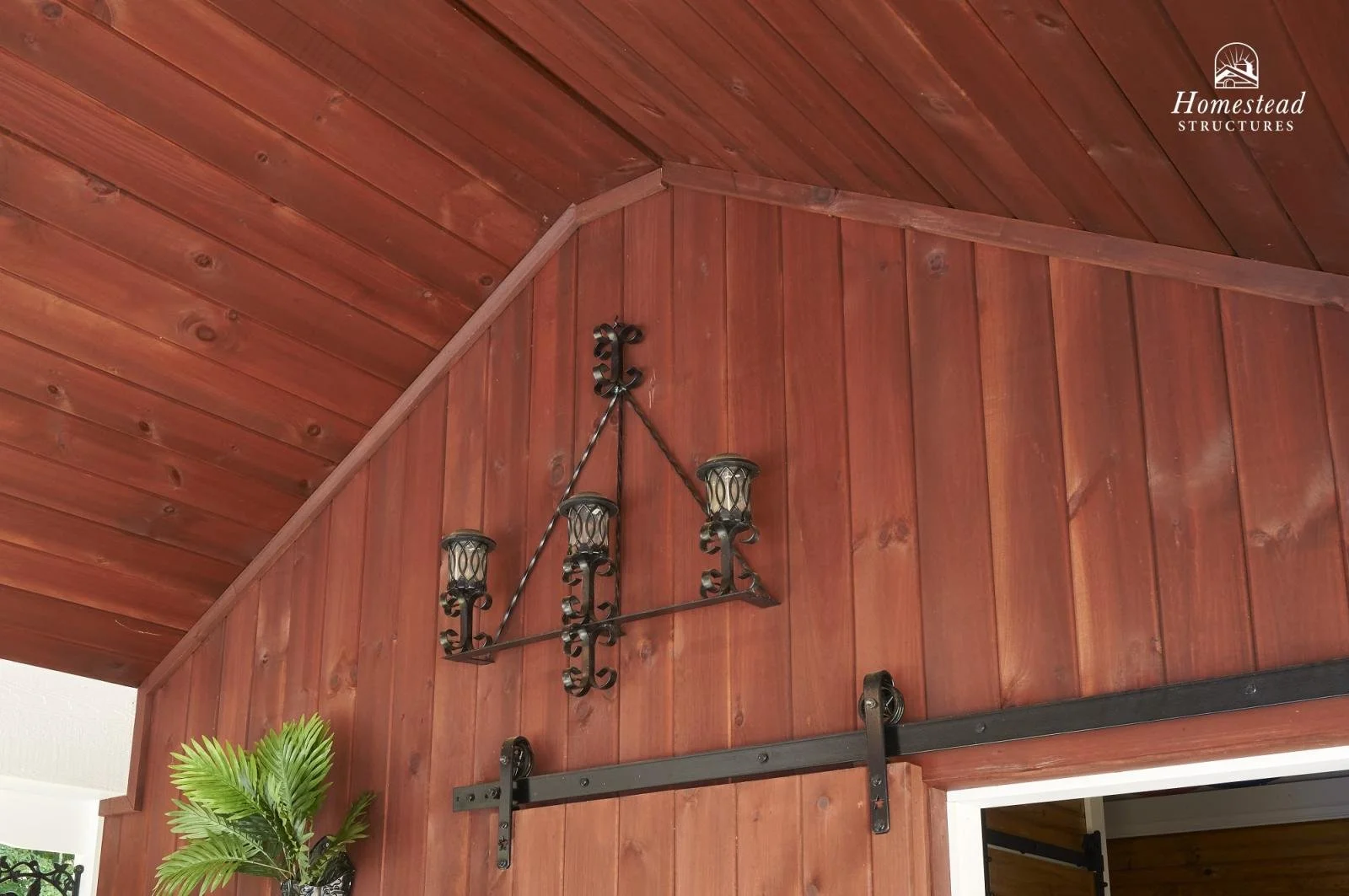 A wrought iron chandelier with four candle-style lights hanging against a wooden-paneled wall and ceiling, with a sliding barn door track below and a potted plant in the bottom left corner.