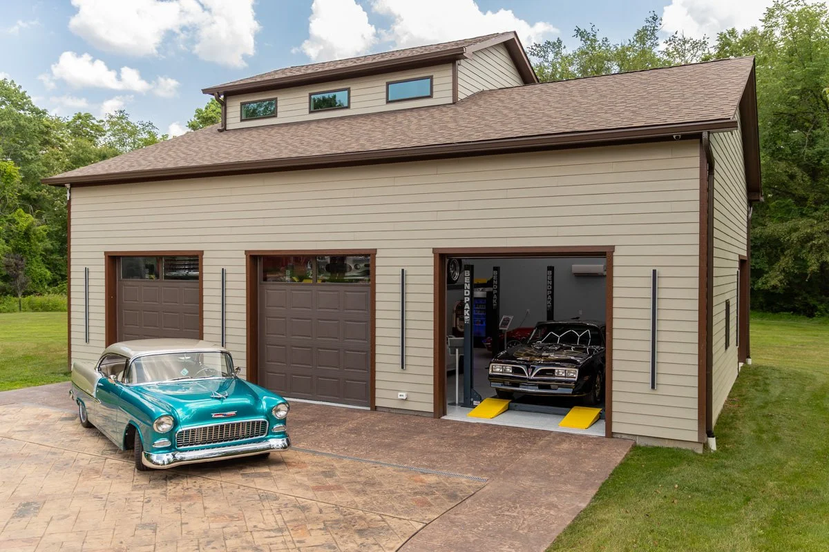 A two-story garage with brown doors and beige siding, containing a museum-quality black car on a lift, and a vintage blue and white classic car parked outside on a pavers driveway, surrounded by green grass and trees.