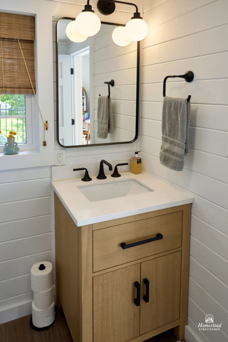 Bathroom vanity with wooden cabinet, white countertop, and black fixtures, a mirror with a black frame, and a wall-mounted towel ring with a gray towel. Light fixtures with round white bulbs are above the mirror, and a window with bamboo blinds and a