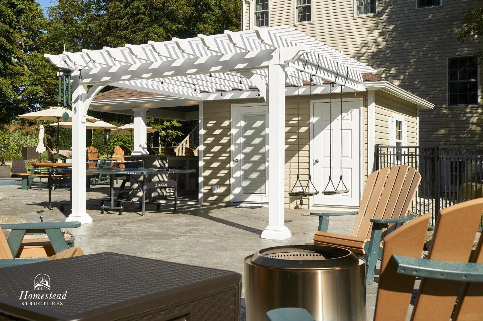 A backyard patio with a white pergola, swing seats, Adirondack chairs, a tiki torch, and a pool area with umbrellas and outdoor furniture. Trees are visible in the background.