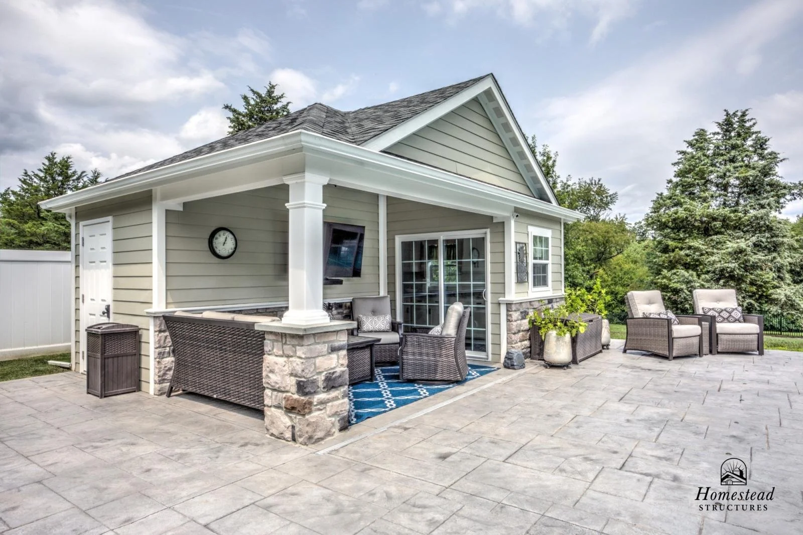 A backyard patio with outdoor seating, a large potted plant, and a house with siding, stone accents, and a sliding glass door, surrounded by trees and a cloudy sky.