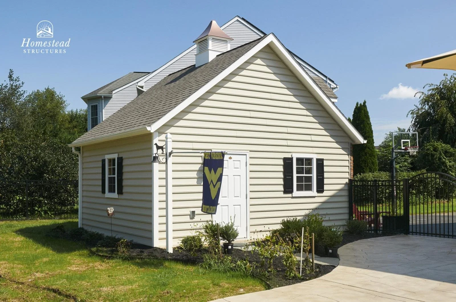 A small beige house with black shutters on two windows, a white door, and a small garden in front, next to a black fence and a basketball hoop in the backyard