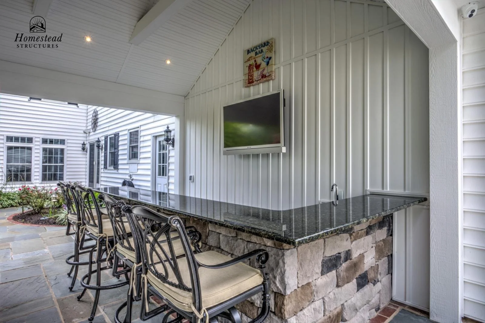 Outdoor covered bar area with a black granite counter, stone base, four bar stools with ornate metal frames and cream cushions, a wall-mounted TV, white siding wall, and garden with flowers in the background.