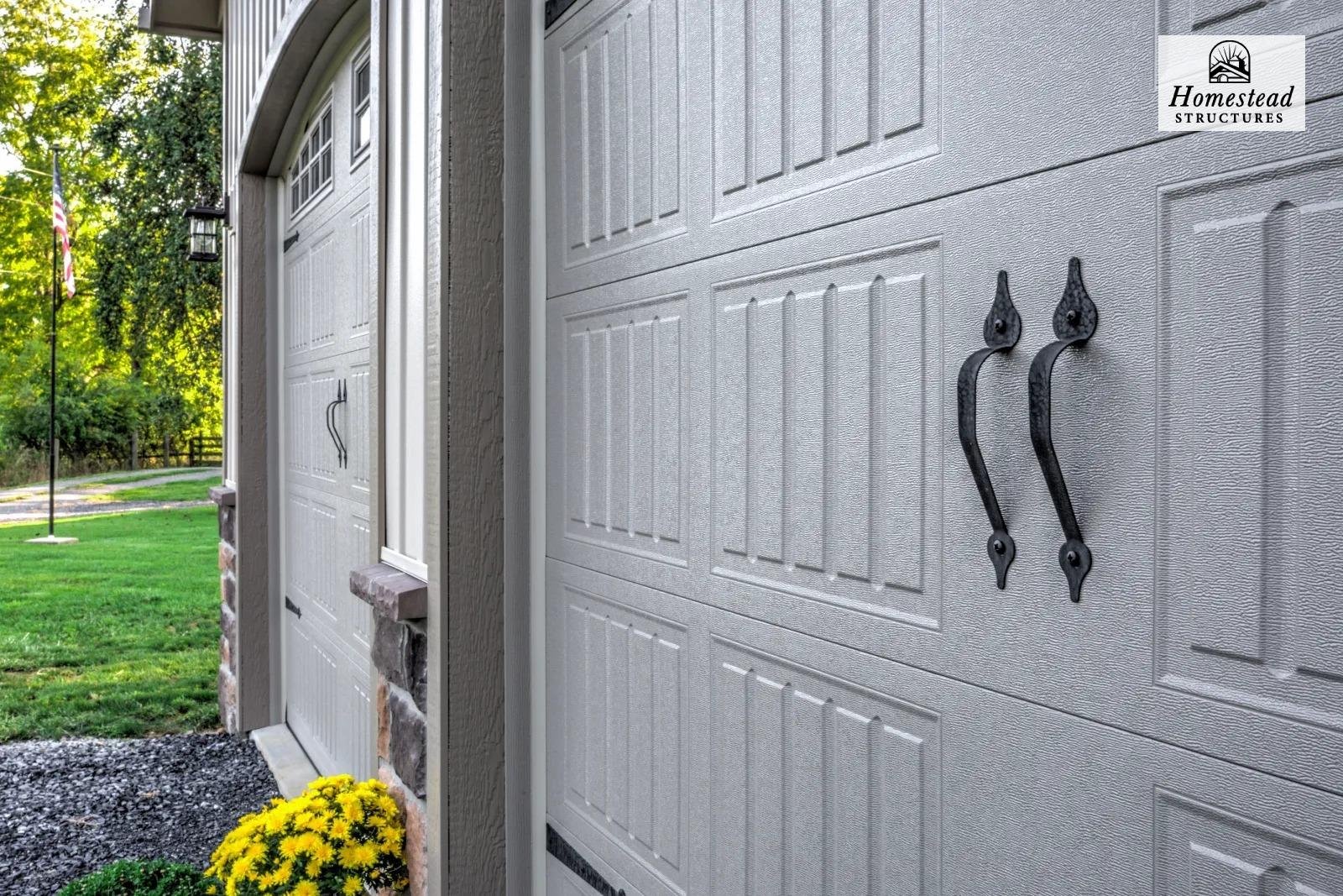 Close-up of a gray garage door with black handles, exterior house wall with a stone lower part, yellow chrysanthemums flower, green grass, sunlight, and a flag in the background.