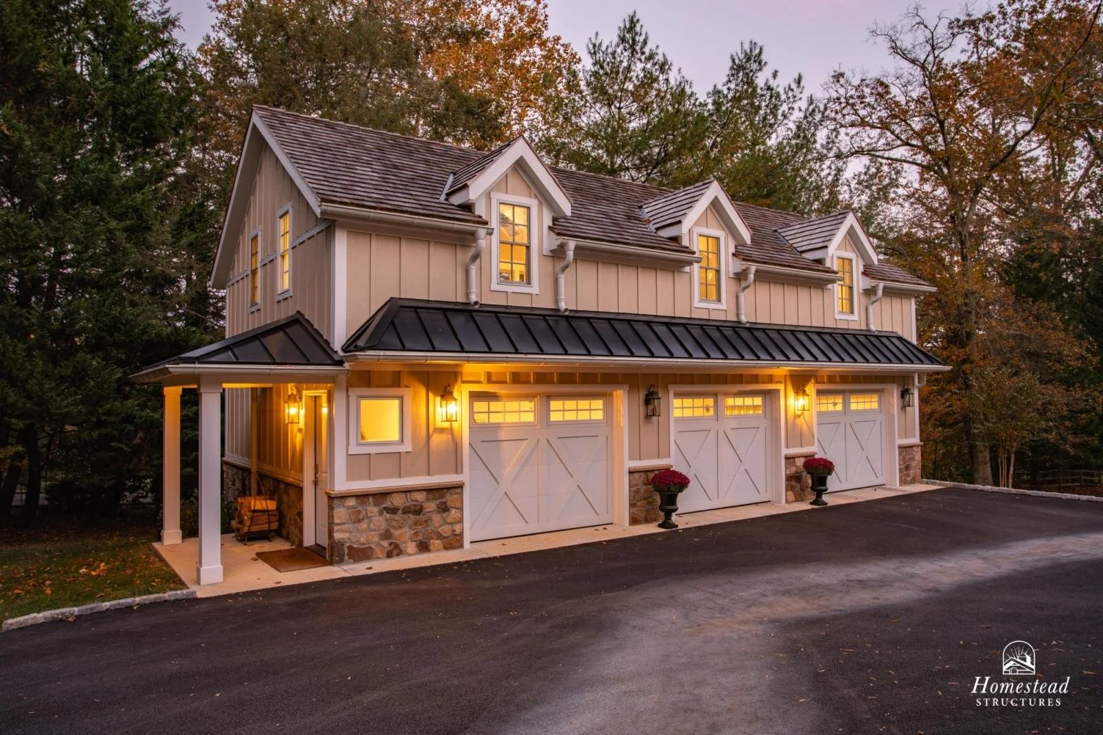 A two-story house with beige exterior walls, white trim, and a black metal roof, illuminated from within, surrounded by trees during dusk with an asphalt driveway in the foreground.