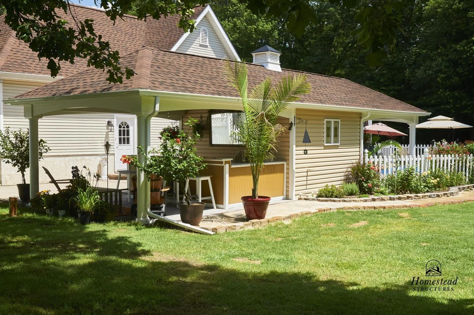 A backyard with a small house or shed featuring a beige exterior with horizontal siding and a brown roof. The patio area has potted plants, outdoor furniture, and a white picket fence with umbrellas. There is a grassy lawn and trees providing shade.