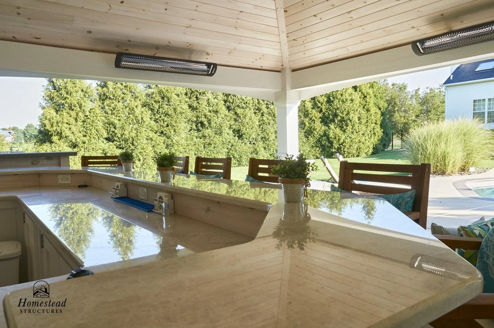 Outdoor covered patio with a marble countertop, wooden chairs, potted plants, and a view of green trees and a house in the background.