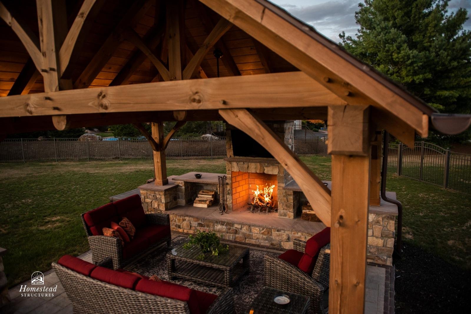 Outdoor seating area with wicker furniture and red cushions under a wooden pergola next to a stone fireplace with a fire burning, in a backyard surrounded by a fence and trees at dusk.