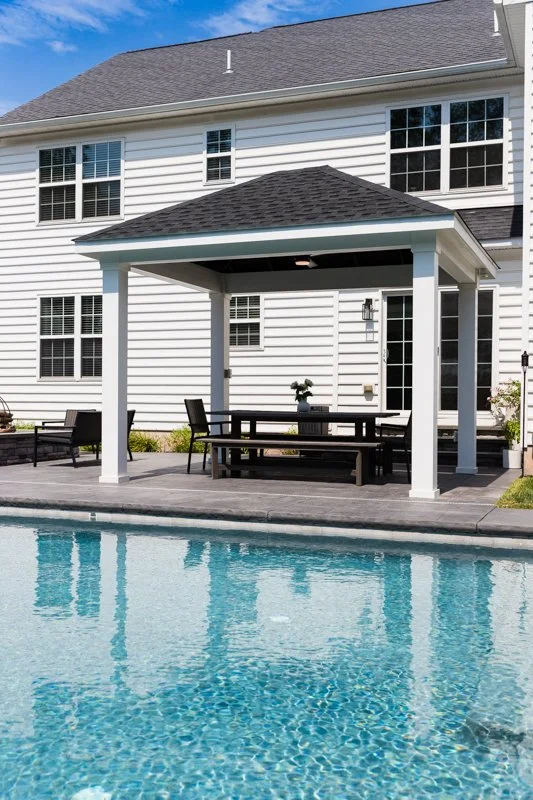 Backyard with a swimming pool, patio furniture under a covered area, and a white house with multiple windows in the background